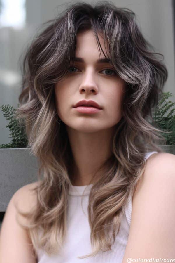 Beauty editorial image of a layered shag haircut with short choppy layers on top and longer textured ends (wolf cut style), dark smokey shadow roots fading into cool ash blonde, soft tousled waves, natural lighting, moody cool tones
